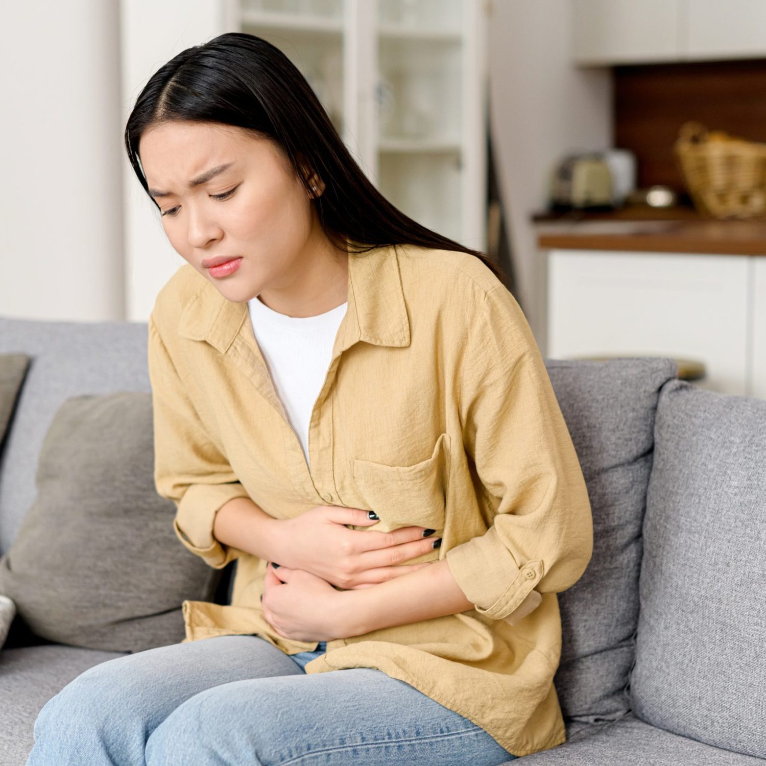 Concerned asian woman suffering from stomach-ache sitting on sofa. Female holding tummy, undergoing belly pain and discomfort, suffer from menstruation